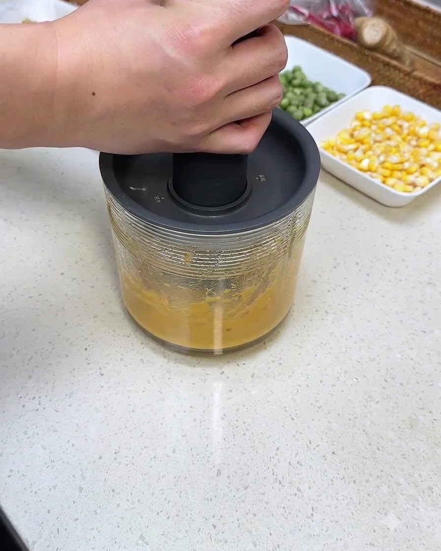 A person pressing down on a manual hand blender unit over a glass container filled with sweet potato puree.