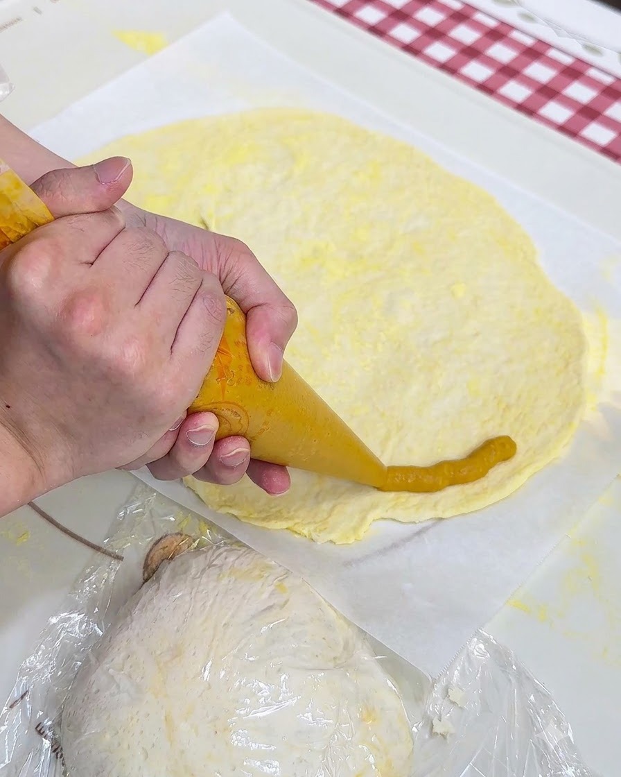 A pair of hands using a piping bag to pipe yellow sweet potato mash around the edge of a flat pizza dough sheet on parchment paper.