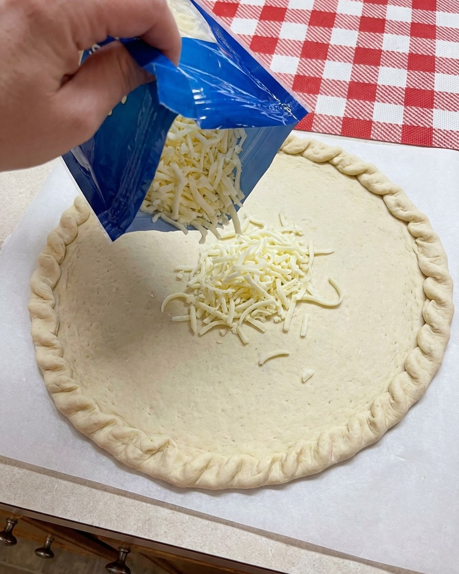 Shredded mozzarella cheese being poured from a blue bag onto the center of a shaped pizza dough base.