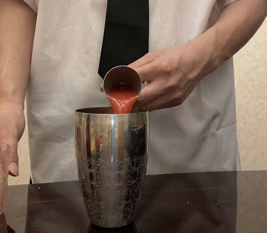 Fresh red tomato juice being poured from a metal jigger into a patterned silver cocktail shaker.