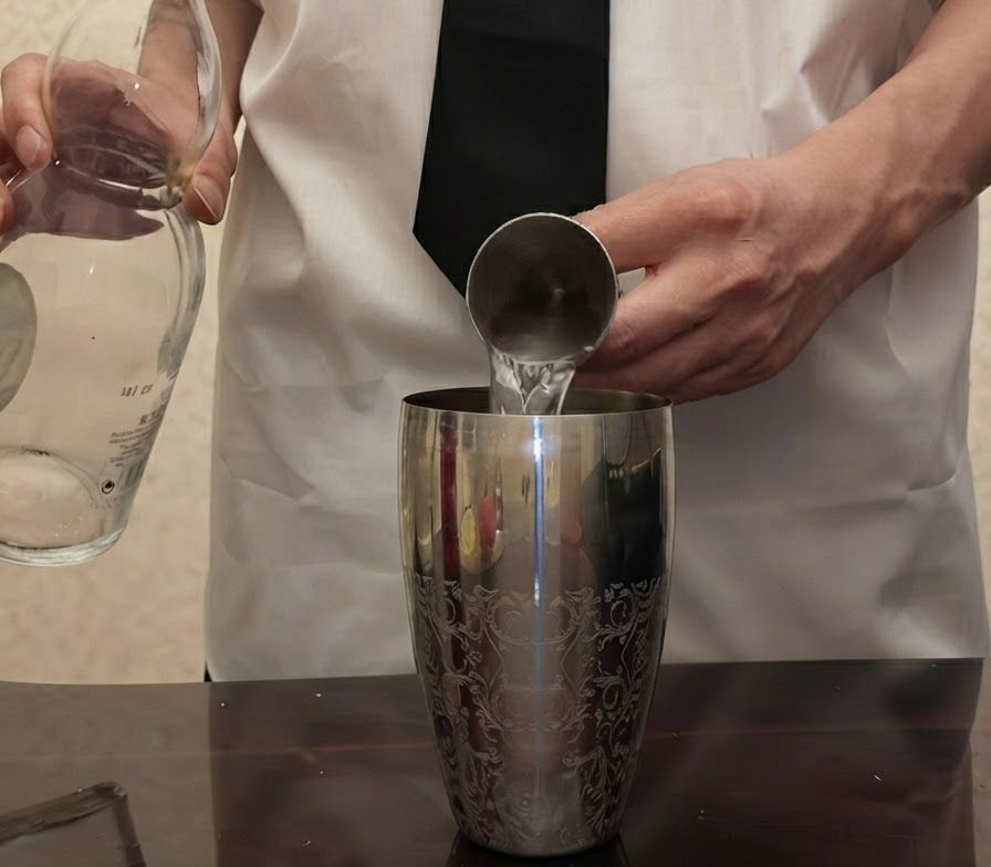 Clear tequila being poured from a glass bottle into a metal jigger held above a silver cocktail shaker.