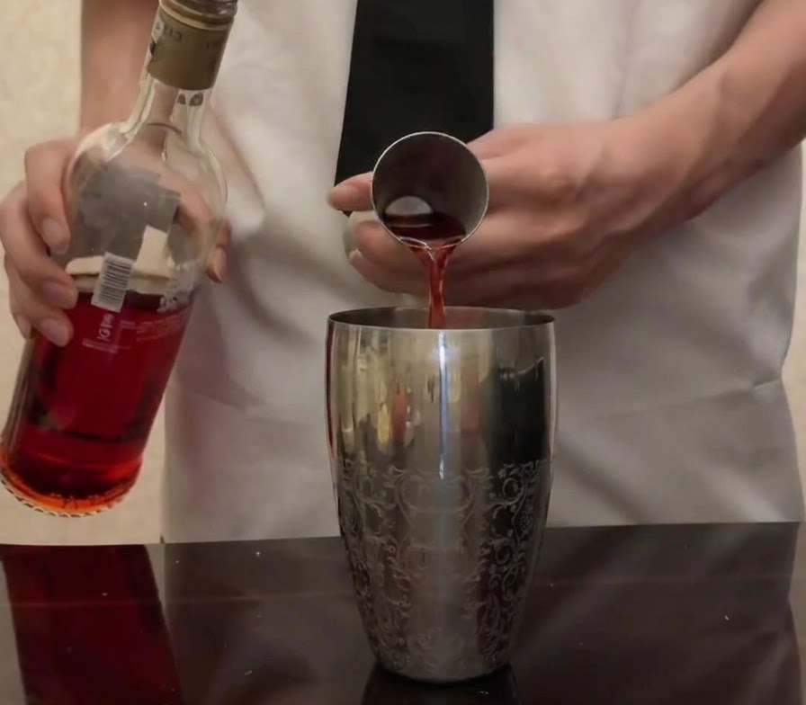 Bright red Campari being poured from its bottle into a metal jigger over a silver cocktail shaker.