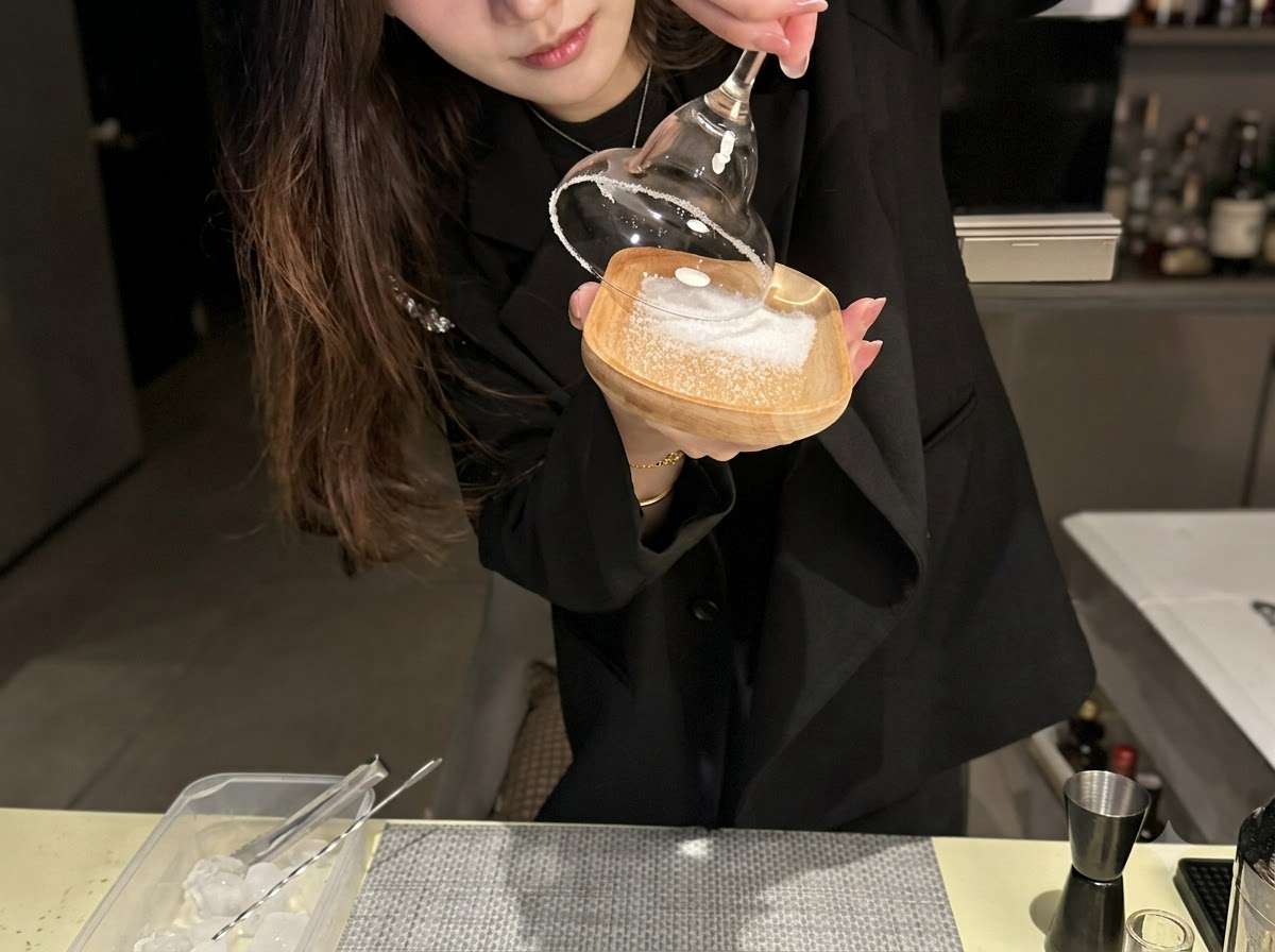 The rim of a cocktail glass being pressed into a layer of sea salt on a small wooden dish.