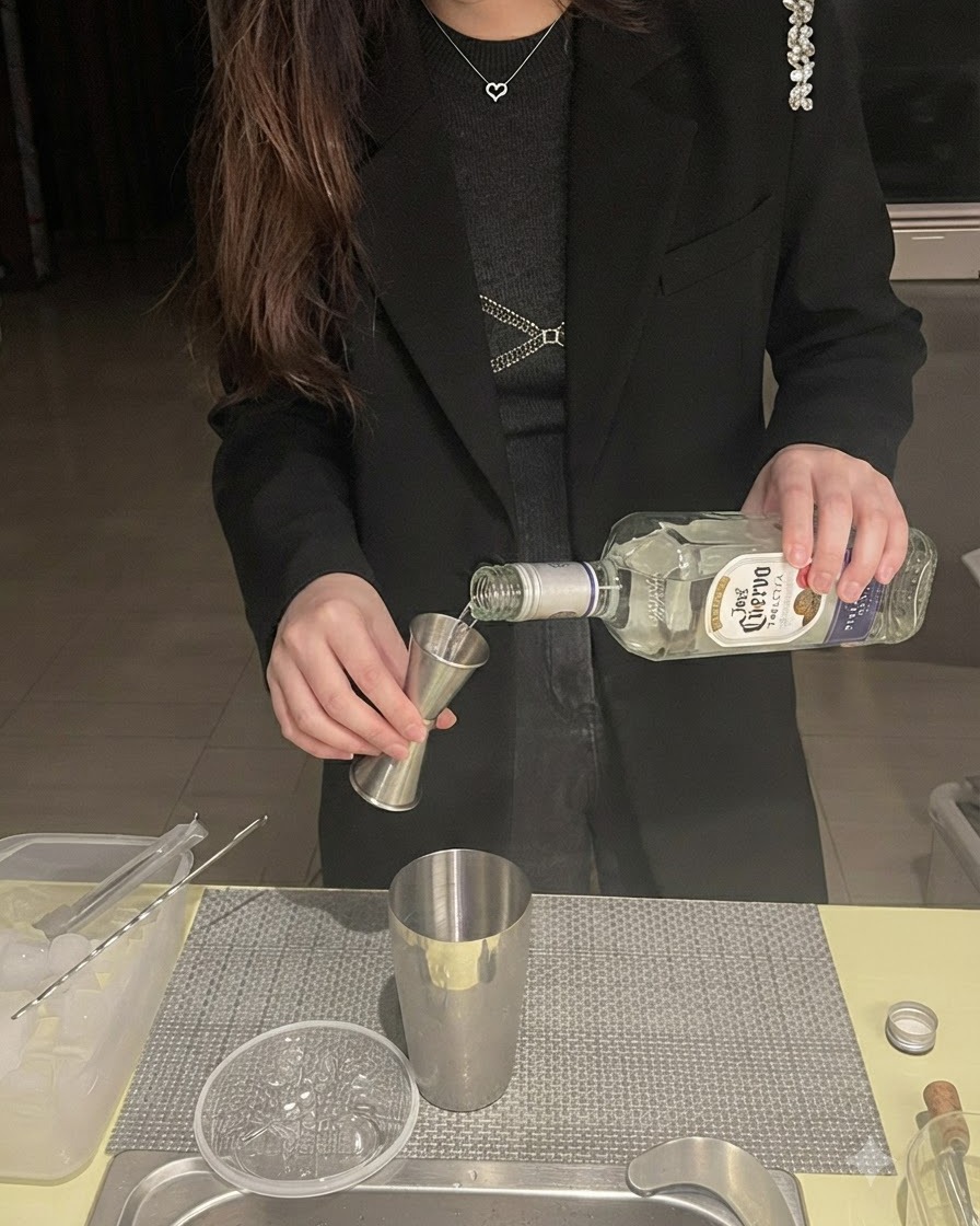 Tequila being poured from a bottle into a jigger positioned over a metal shaker.
