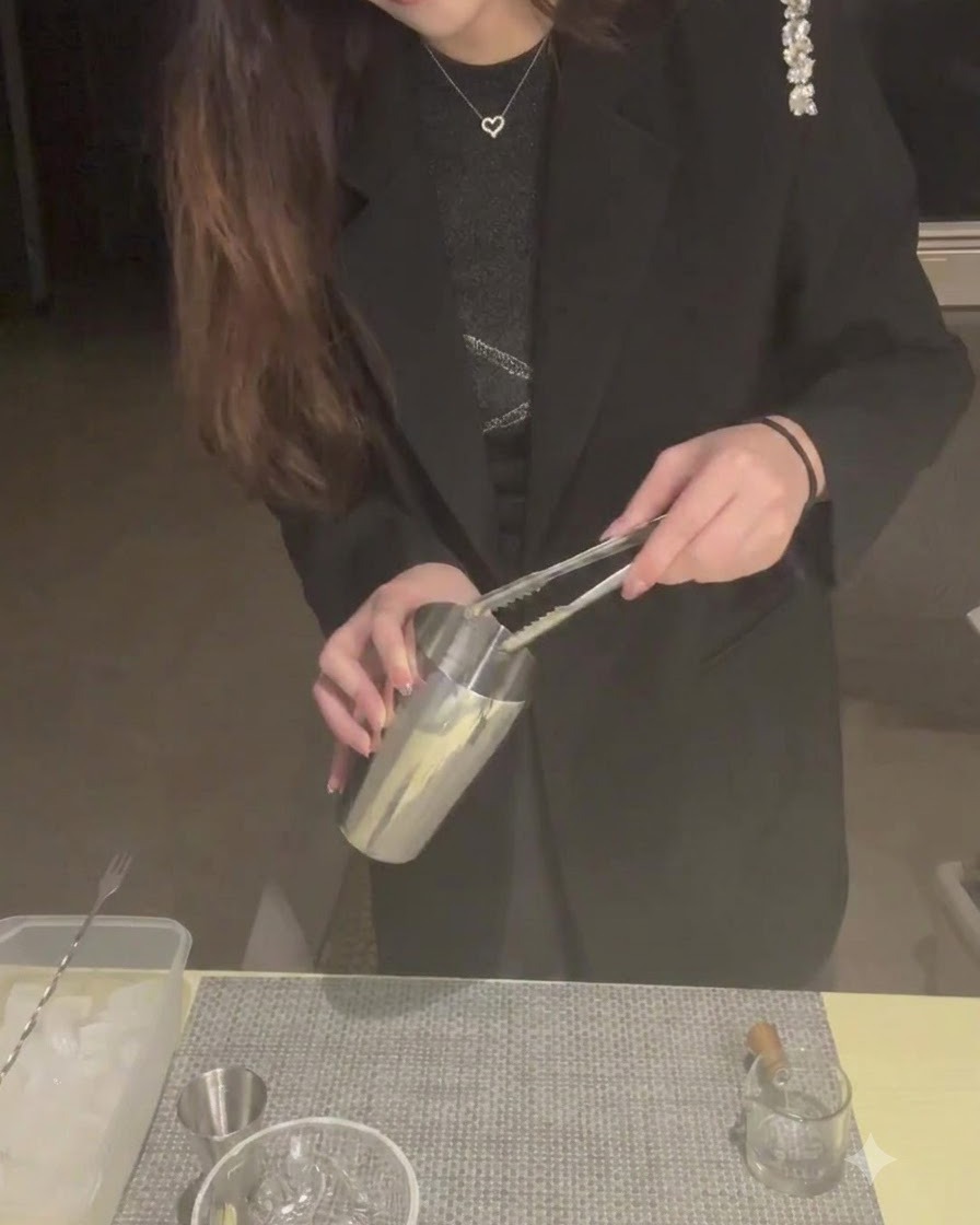 A person using tongs to place ice cubes into a metal cocktail shaker tin.