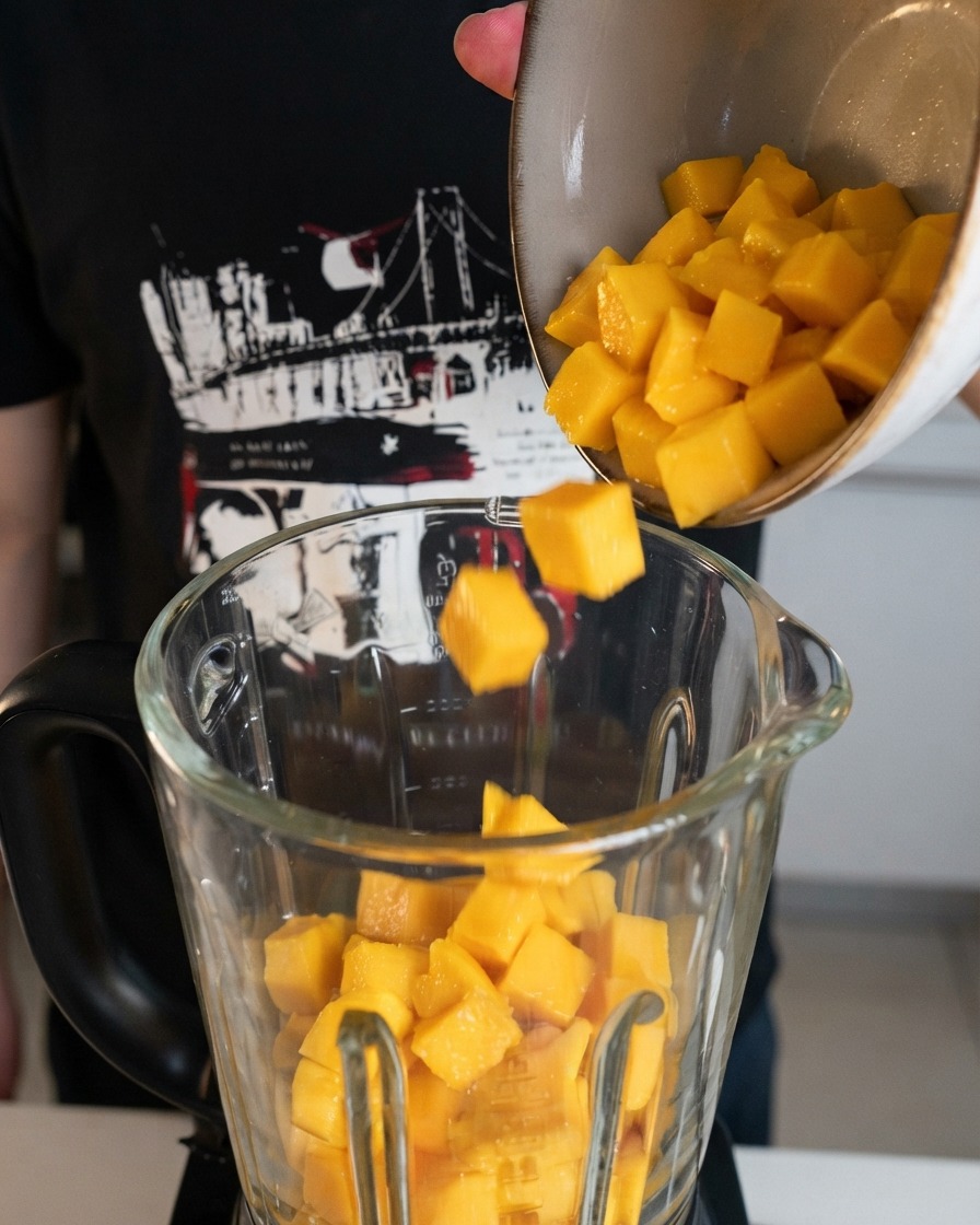 Freshly cut yellow mango cubes being poured from a bowl into a clear glass blender jar.