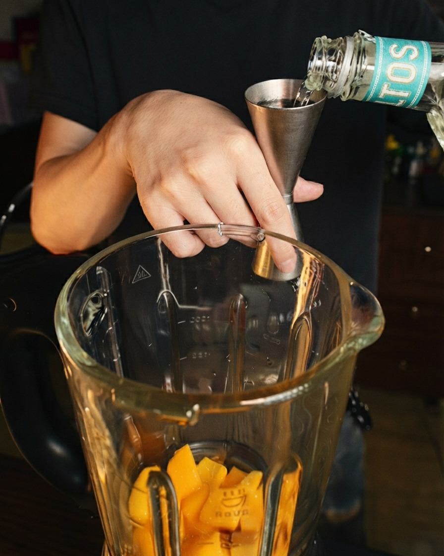 Clear liquor being poured from a stainless steel jigger into a glass blender pitcher over fresh mango pieces.