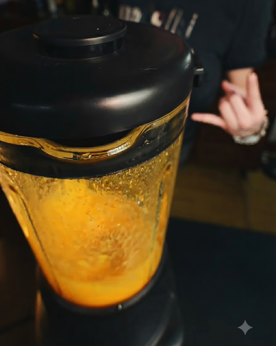 A blender operating with a bright orange mango mixture inside, with a hand making a gesture in the background.