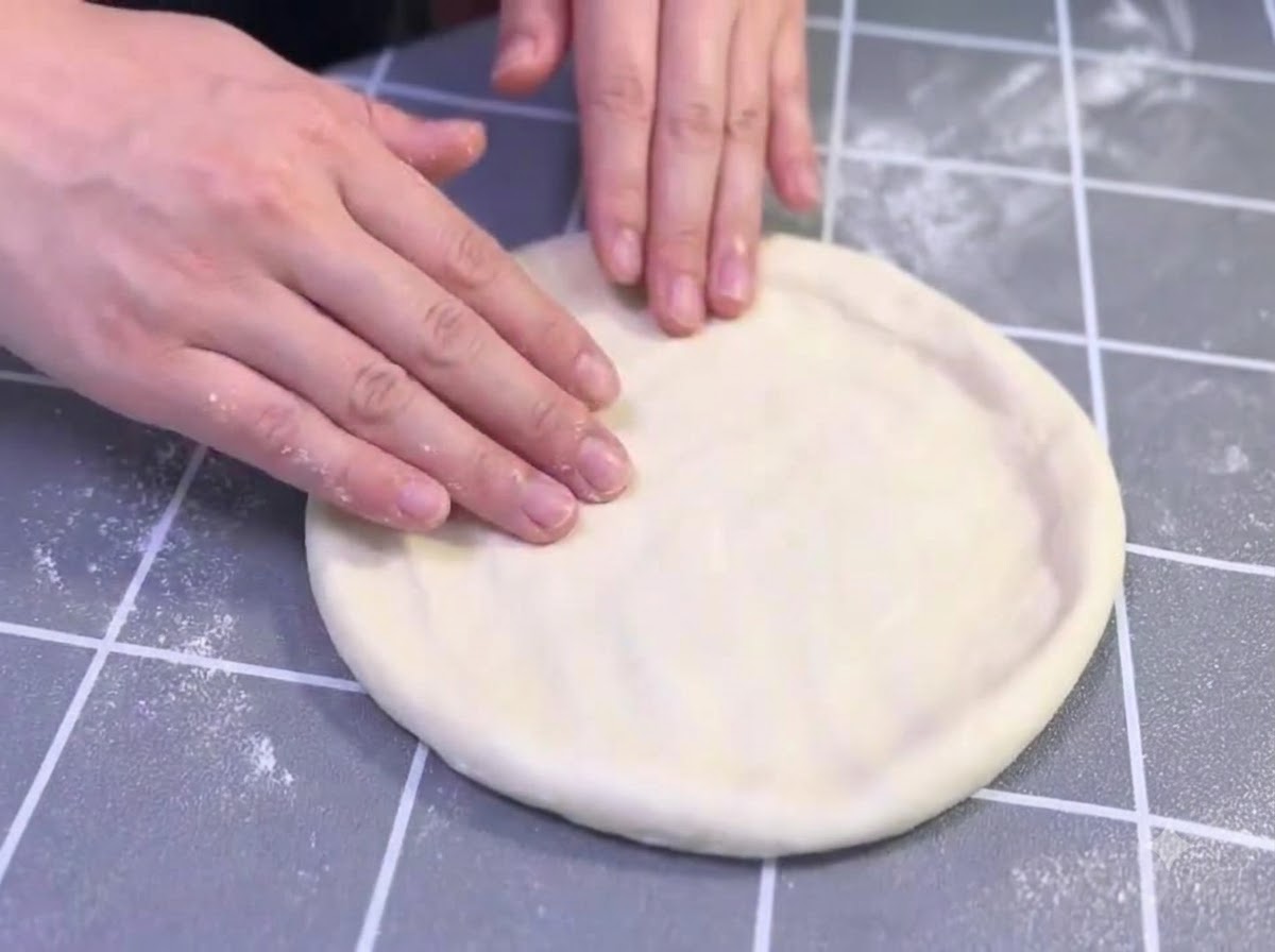 Hands gently stretching and shaping an oiled pizza dough round on a grey gridded surface.