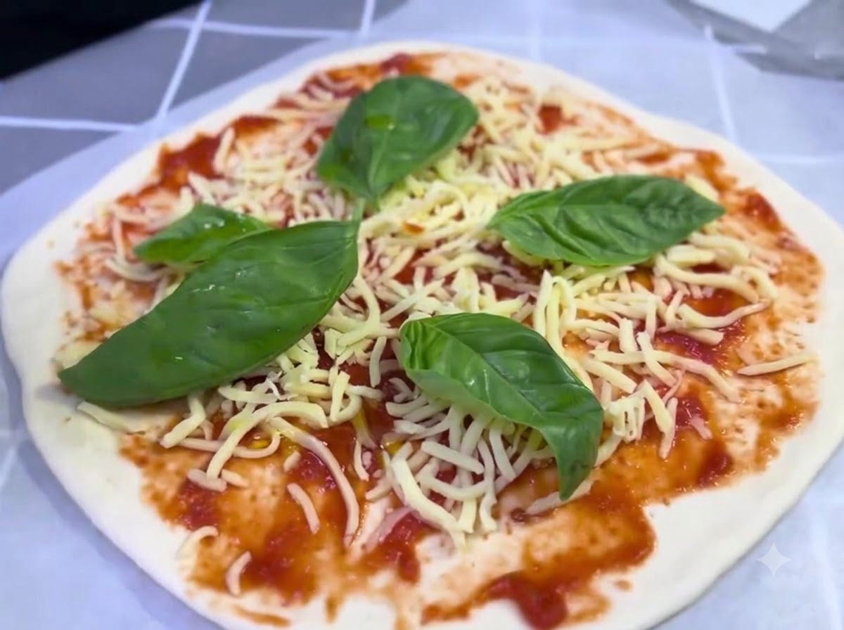 An assembled pizza topped with cheese and basil leaves receiving a light drizzle of olive oil.