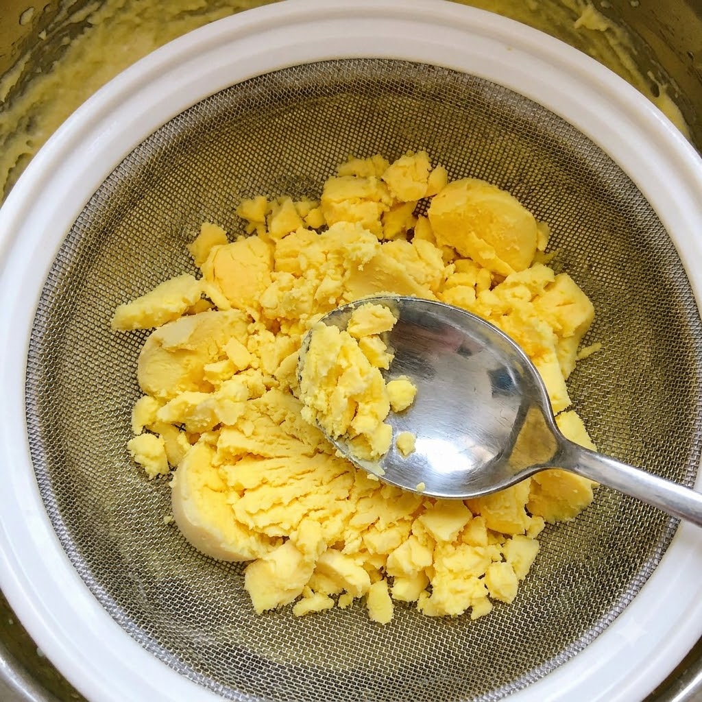 Sieving boiled egg yolks through a wire mesh strainer using a metal spoon.