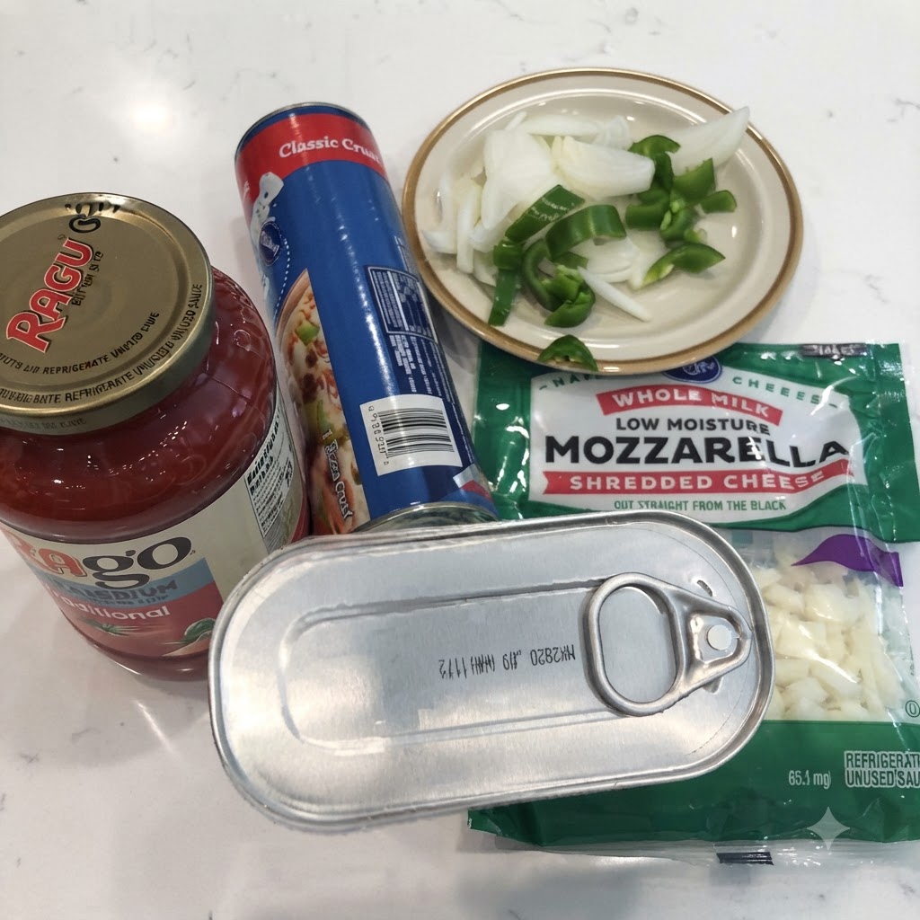 A can of anchovies, jar of marinara sauce, canned pizza dough, shredded mozzarella, and sliced onions and peppers on a white countertop.