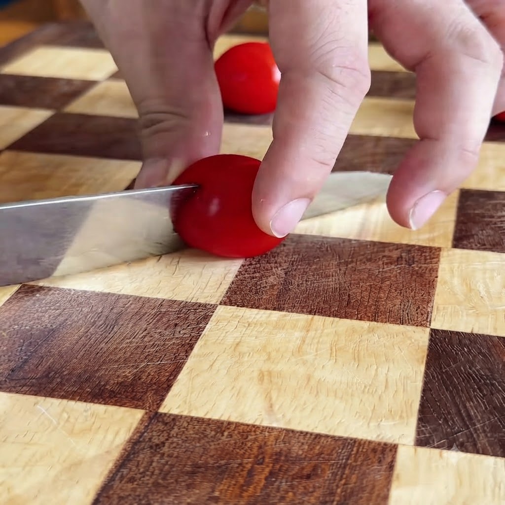 Hands slicing a red cherry tomato in half with a knife on a checkered cutting board.