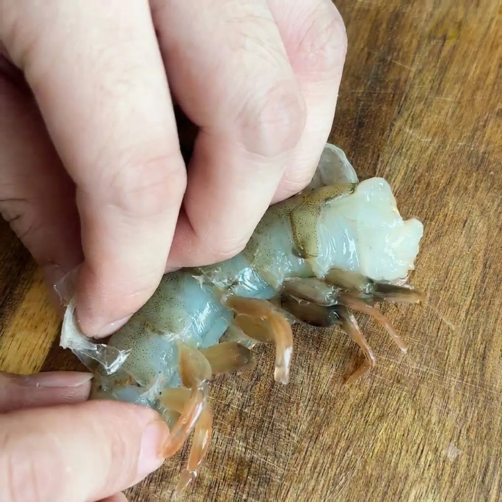 Fingers pulling the translucent shell off a raw shrimp on a wooden surface.