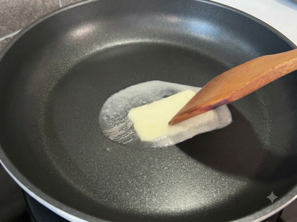A wooden spatula pressing lightly on a square pat of butter melting in a black frying pan.