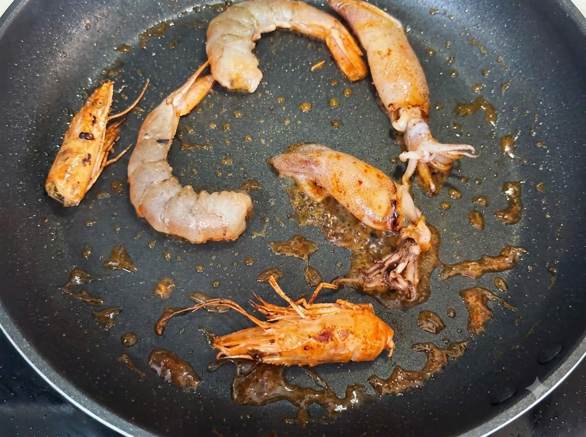 Shrimp, whole shrimp heads, and squid pieces frying in a pan, seasoned with dark specks of black pepper.