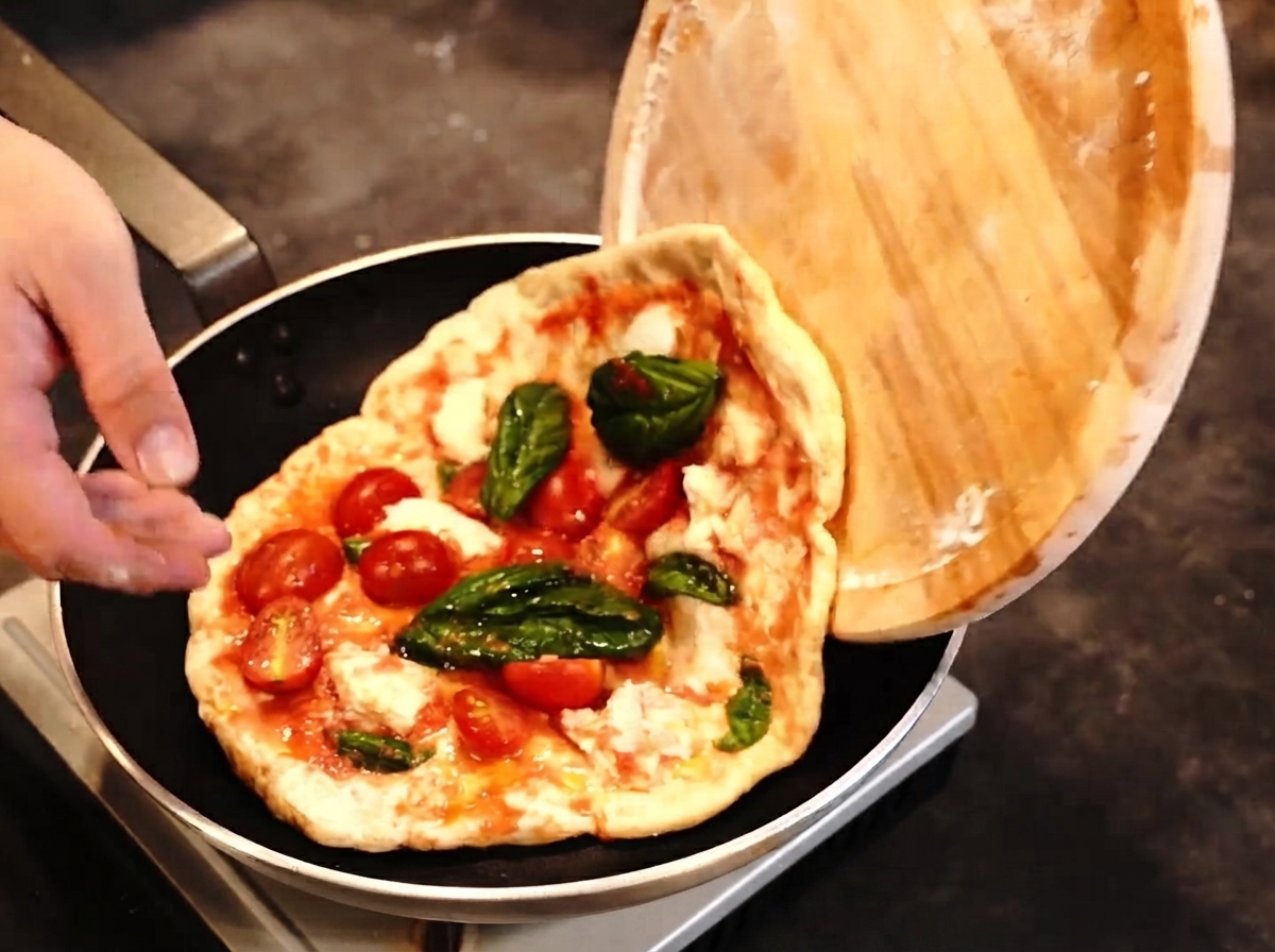 A pizza being transferred from a wooden peel into a frying pan while a blowtorch is ready nearby.