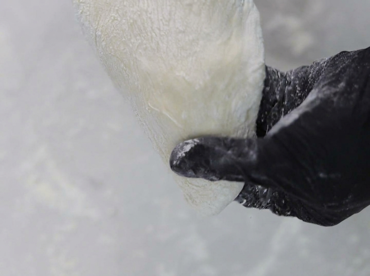 Hands wearing black gloves stretching and shaping pizza dough on a floured surface.