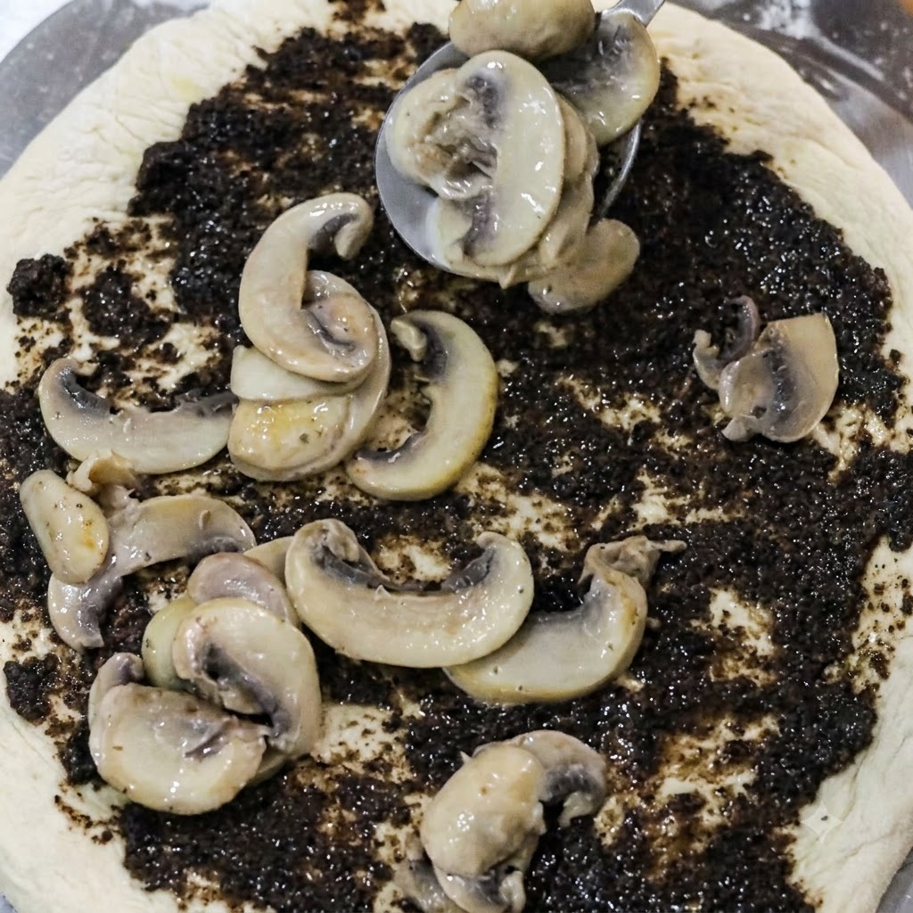 Spoon placing cooked mushroom slices onto a pizza base coated with black truffle paste.