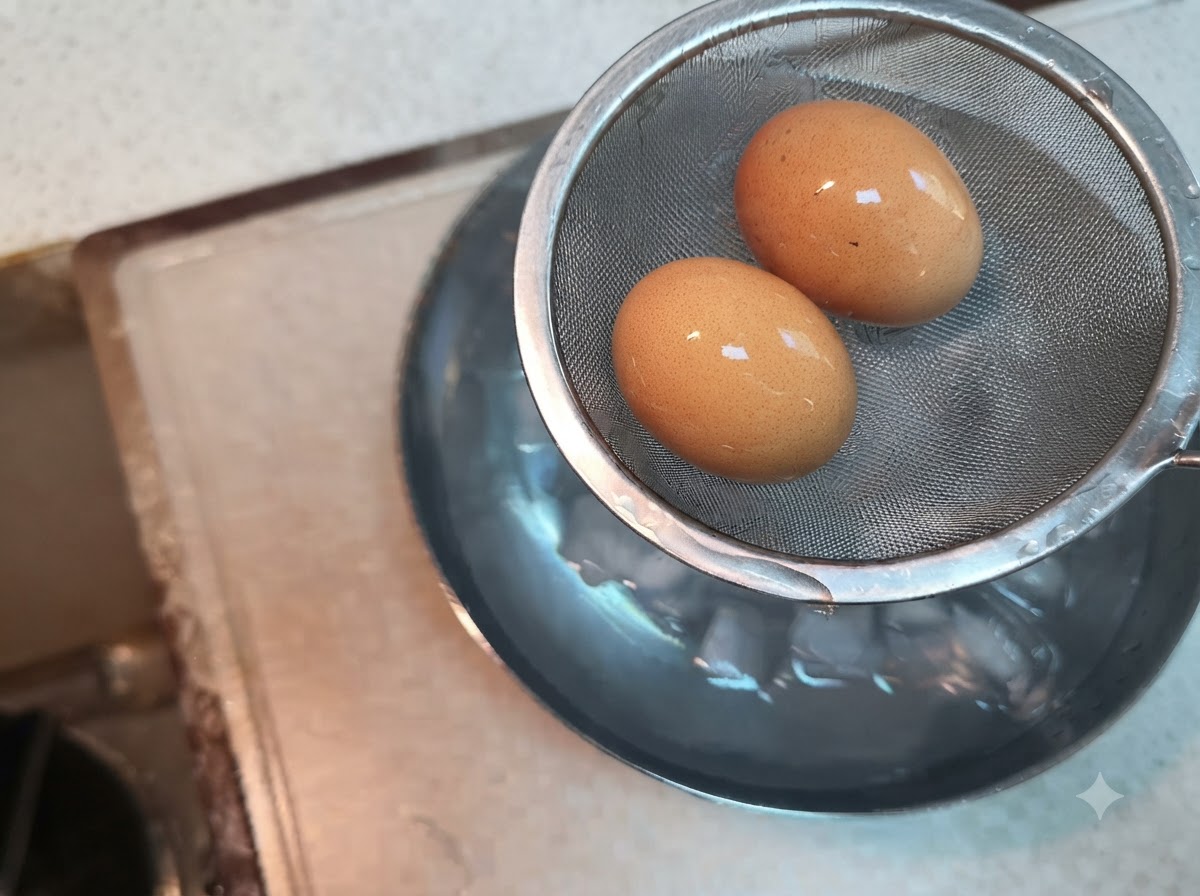 Two boiled eggs resting inside a wire mesh strainer held above a bowl of ice water.