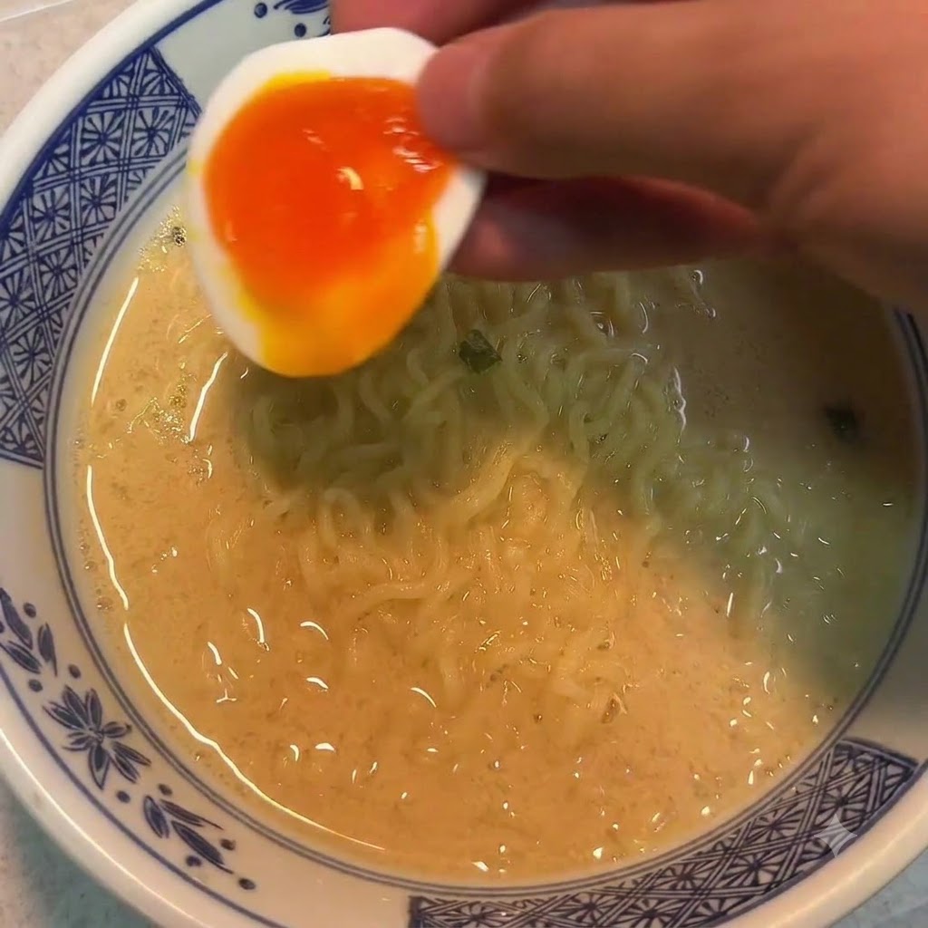 A hand holding half of a soft-boiled egg over a bowl of noodles.