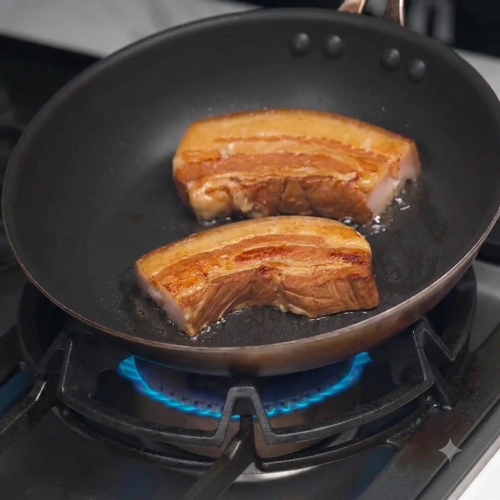 Thick slices of braised pork belly crisping and browning in a hot black frying pan on a gas stove.