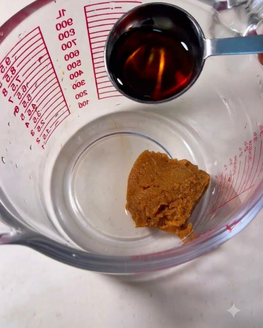 A measuring cup containing a dollop of brown miso paste with a spoon pouring mirin into it.