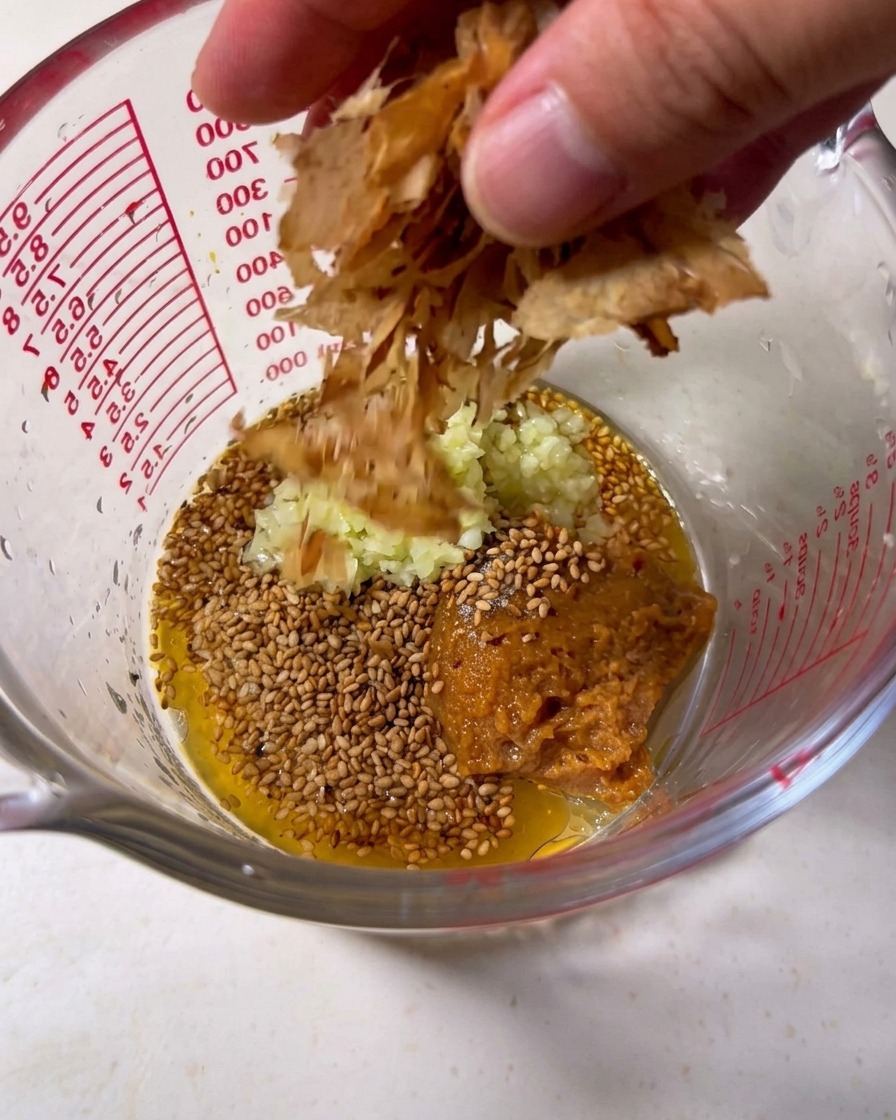 Tonkotsu broth being poured into a measuring cup filled with sesame seeds, minced garlic, and bonito flakes.