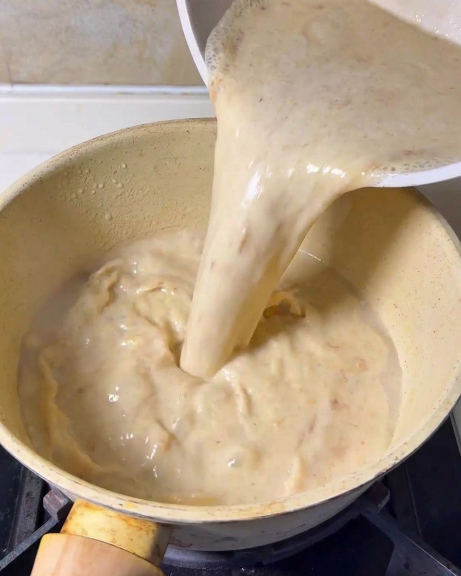 Blended creamy soup base being poured from a container into a yellow cooking pot on a stove.