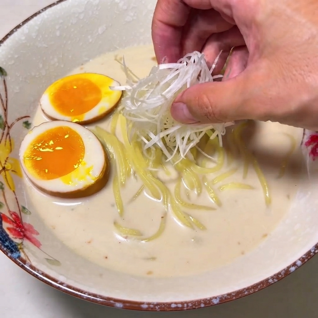 A hand placing a mound of shredded white green onions onto a bowl of ramen.