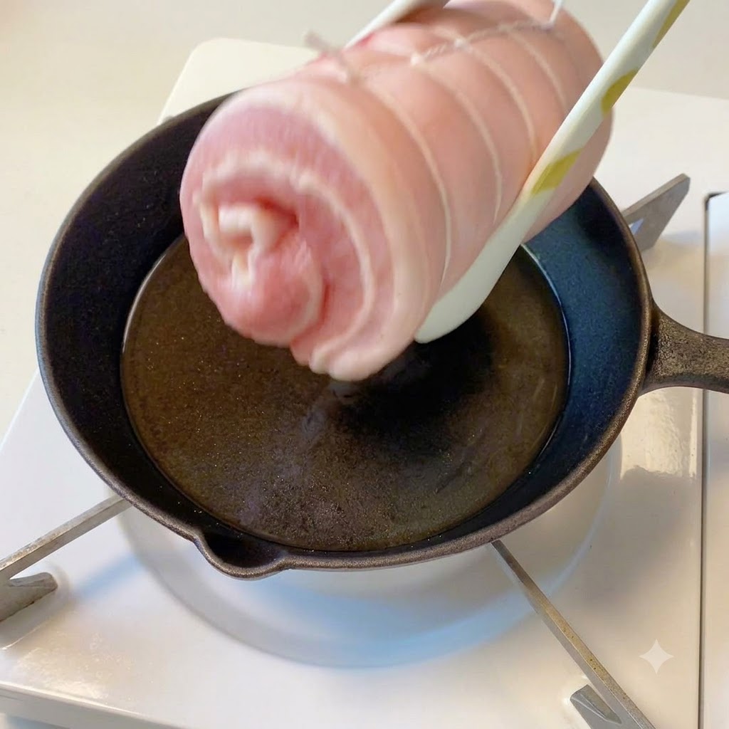 The tied pork belly cylinder being held with tongs over a hot skillet with oil.