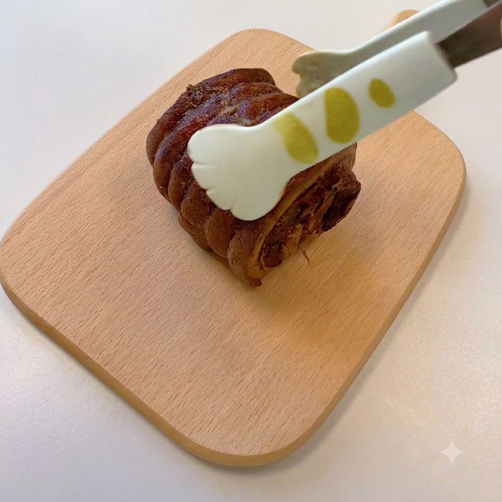 A pair of tongs holding a cooked, rolled chashu pork piece resting on a wooden cutting board.