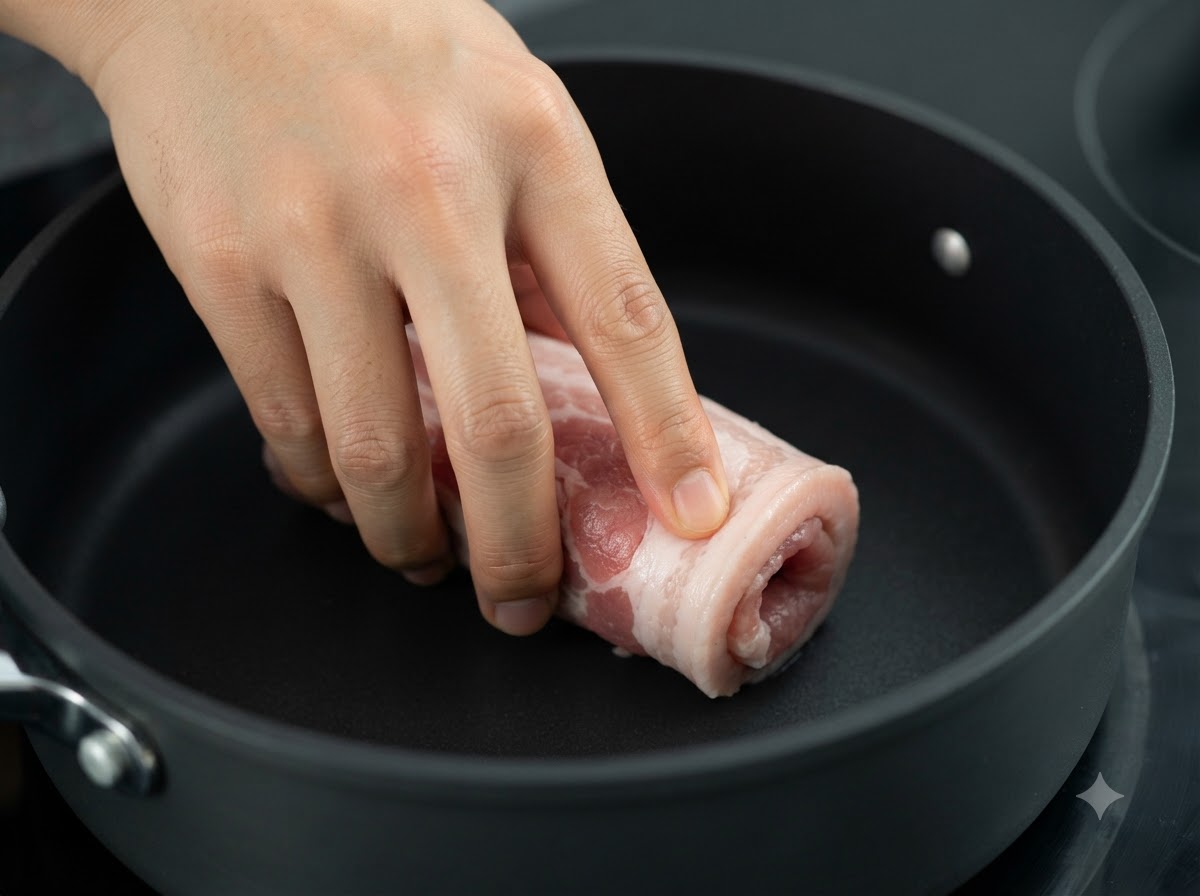 The tied pork belly roll being seared in a dark pan over heat.