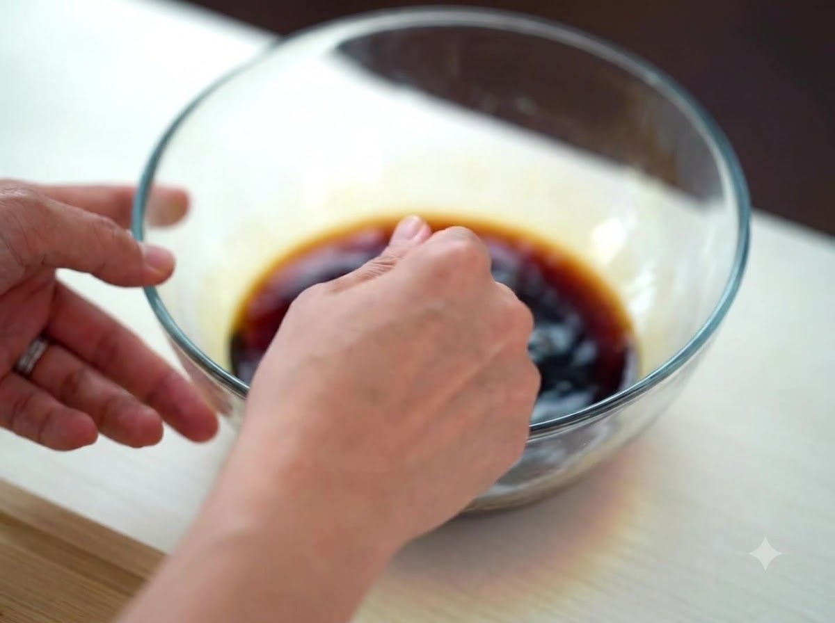 A hand mixing a dark soy-based sauce in a clear glass bowl.