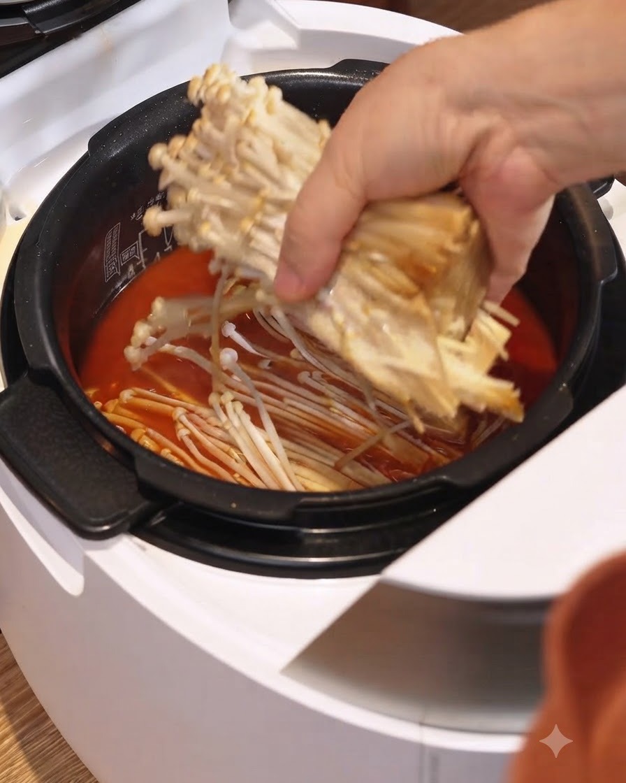 A bunch of enoki mushrooms being placed into the red stew broth inside a white electric rice cooker.