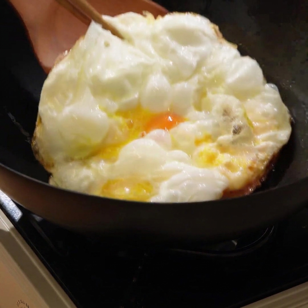 A large golden-brown fried egg being flipped in a hot wok with a wooden spatula.