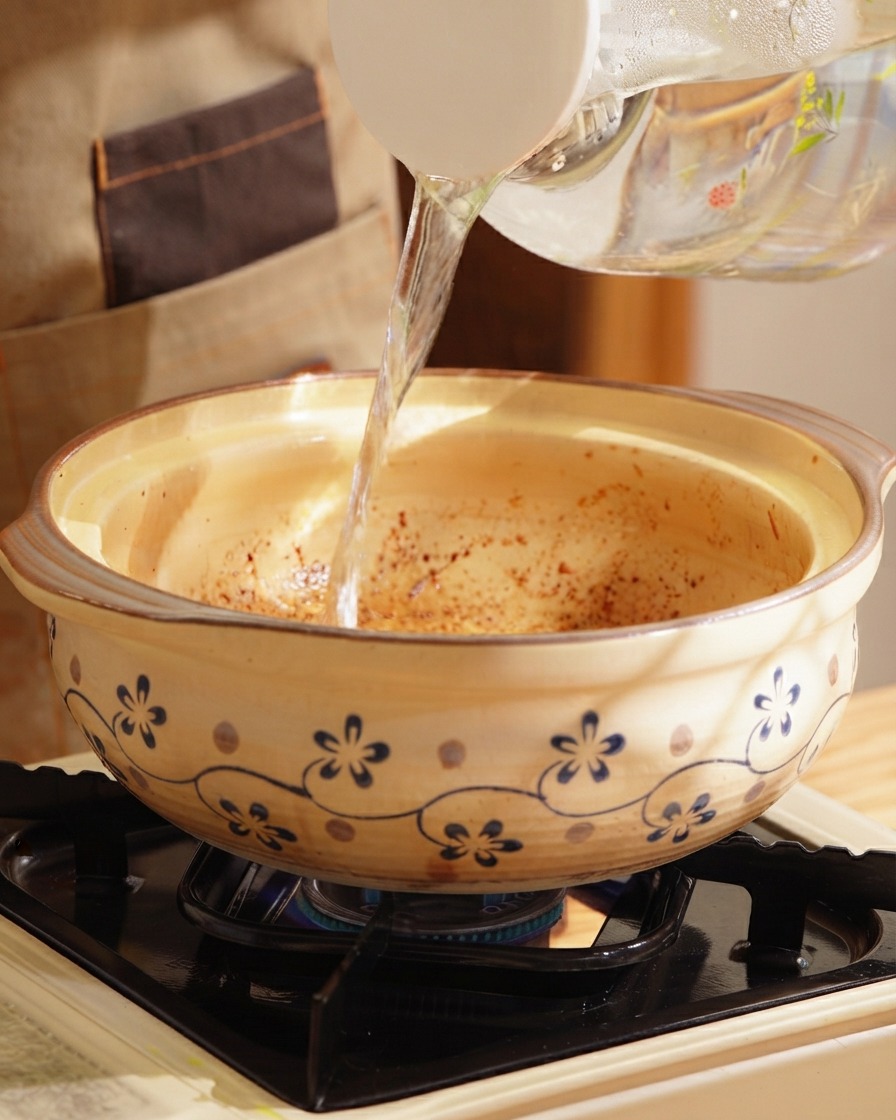 Clear hot water pouring from a pitcher into a decorated clay pot on a stovetop.