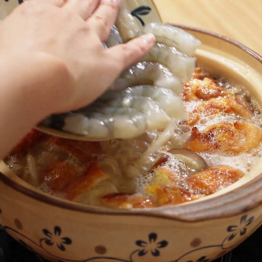 Raw peeled shrimp being added into a bubbling clay pot filled with soup and fried egg pieces.