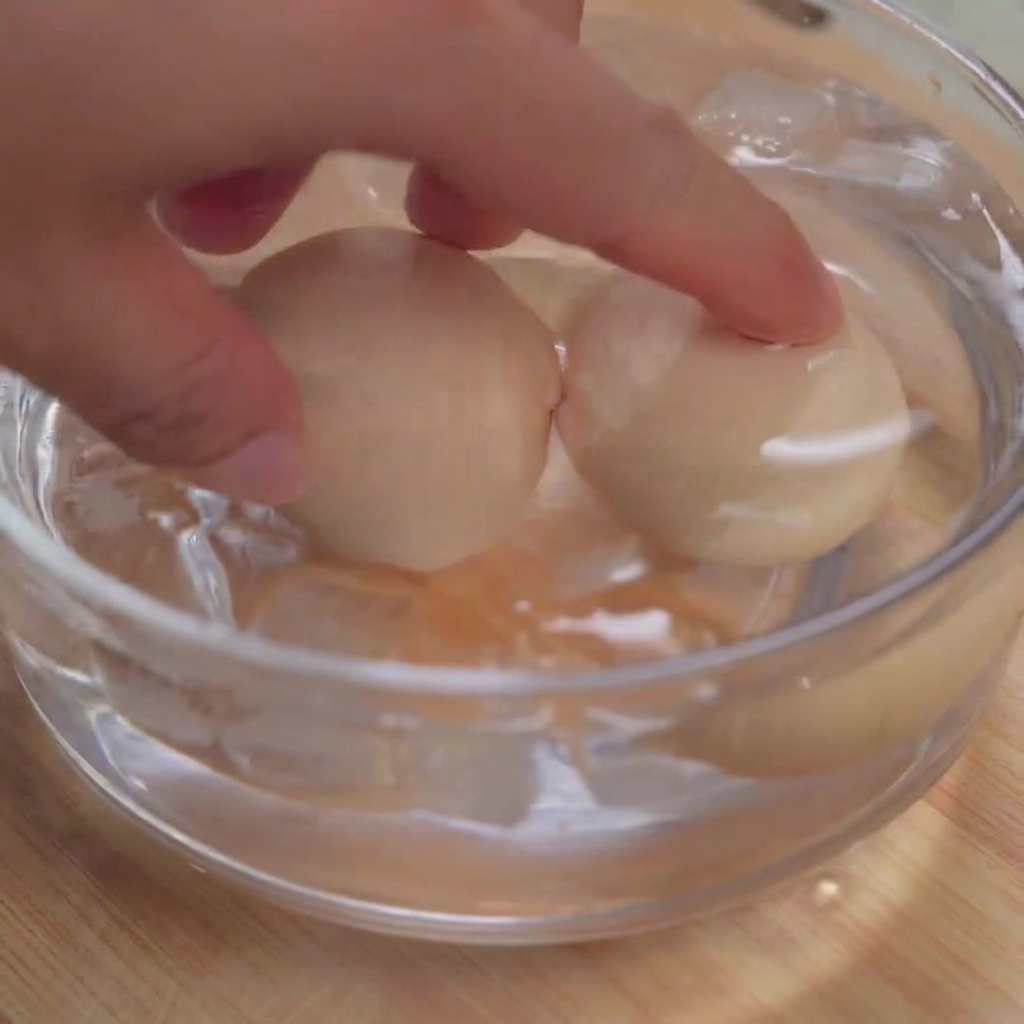 Two soft-boiled eggs being submerged into a clear glass bowl filled with ice cubes and cold water.