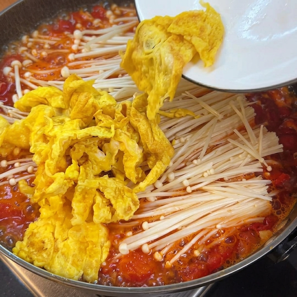 Tender tofu cubes being added into the pot containing tomato broth, mushrooms, and eggs.