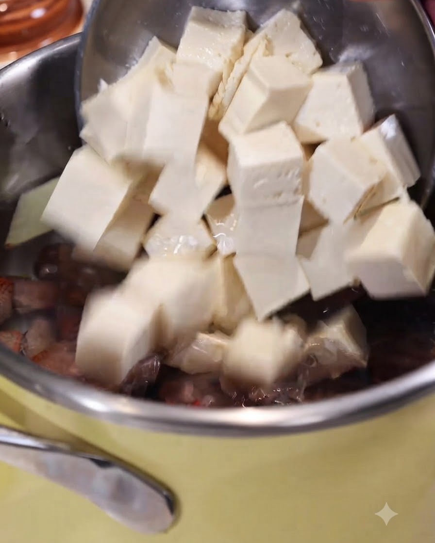 Cubed tofu being slid off a slotted spoon into a pot of boiling water with ginger and clams.