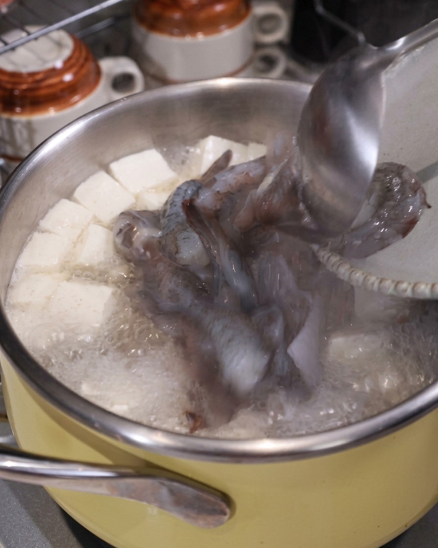 A ladle adding raw squid and shrimp pieces into a boiling pot of soup with tofu and pork.