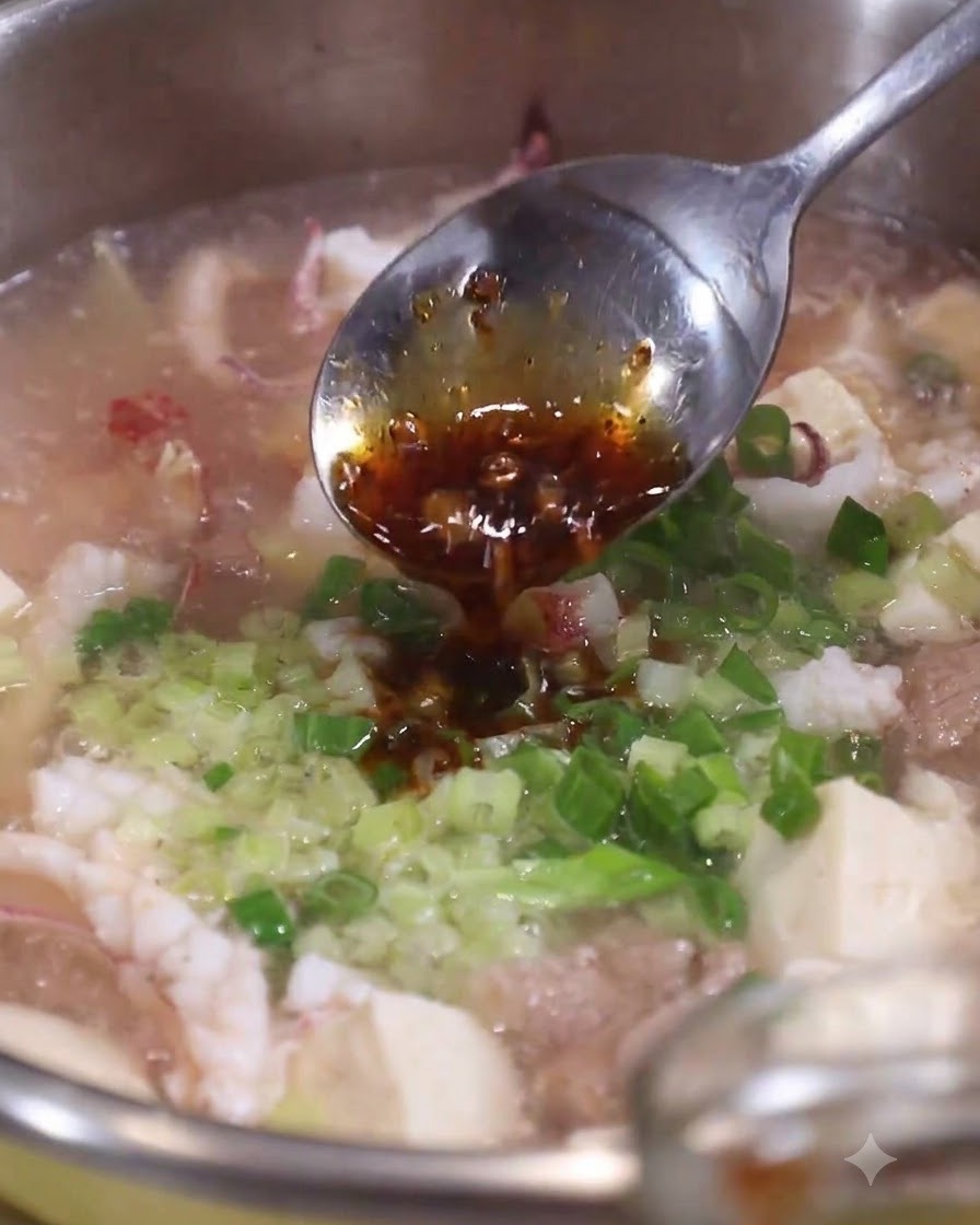 A spoonful of dark scallion oil being drizzled over fresh chopped green onions and celery in a pot of hot seafood soup.
