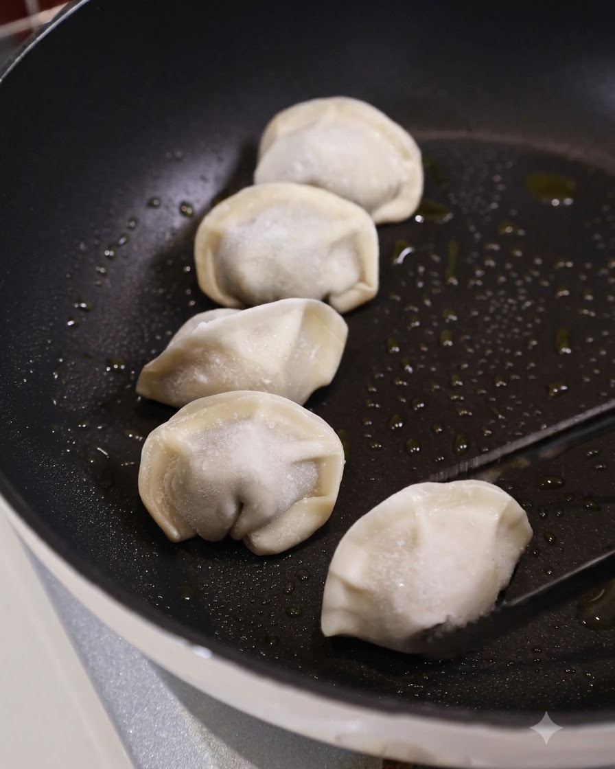 Kitchen tongs placing a raw frozen dumpling into a dark, oiled flat pan alongside several other dumplings.