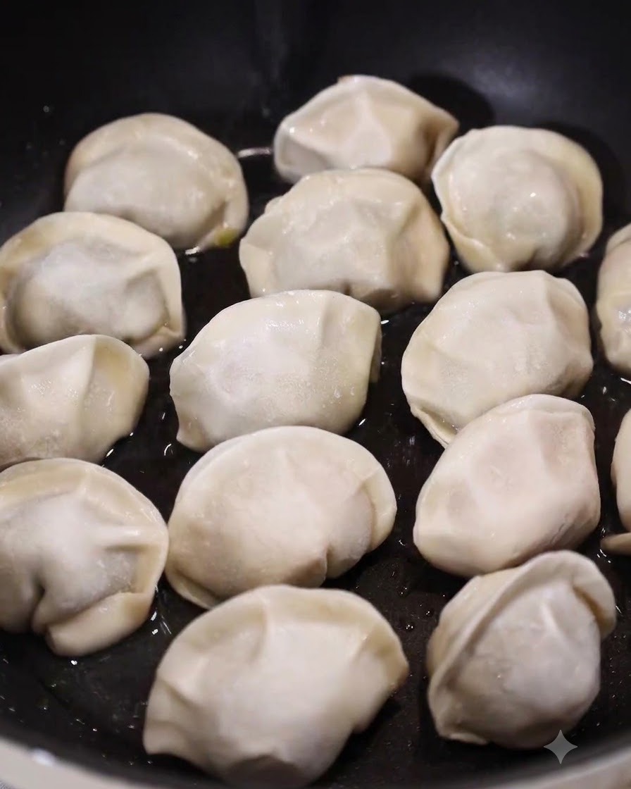 A flat pan filled with raw dumplings resting in a shallow layer of bubbling water and oil.
