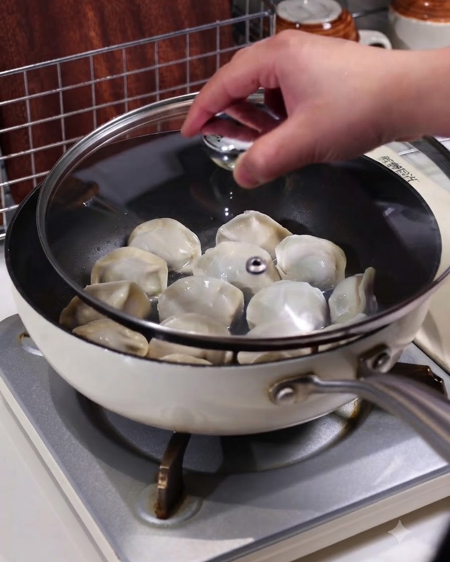 A hand placing a transparent glass lid onto a pan containing steaming dumplings on a stovetop.