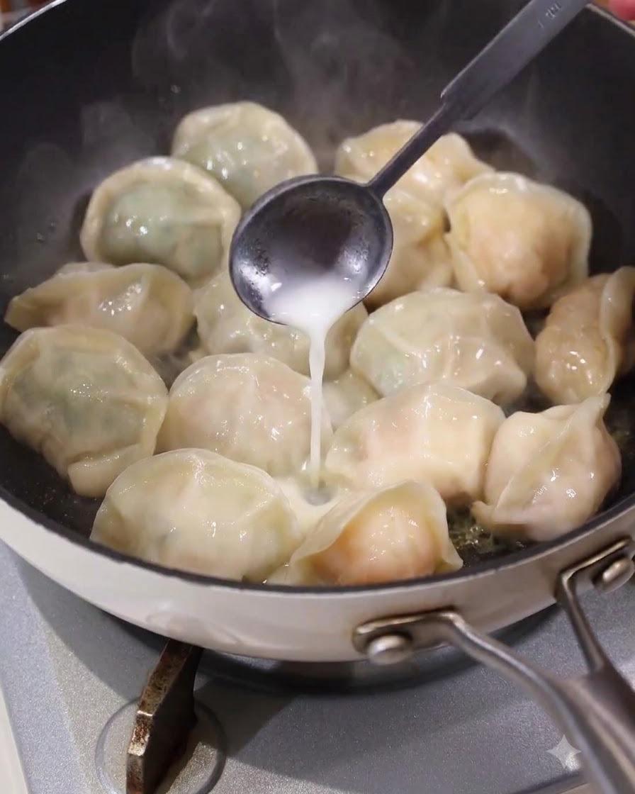 A spoon pouring a white liquid slurry evenly into the spaces between dumplings in a hot frying pan.