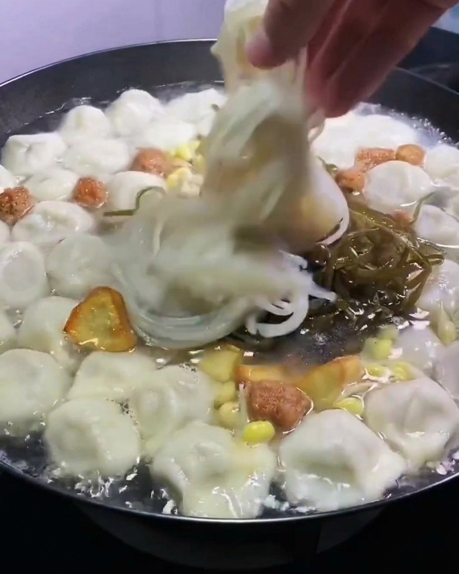 A hand placing shredded green kelp and translucent glass noodles into a pot of boiling dumplings and broth.