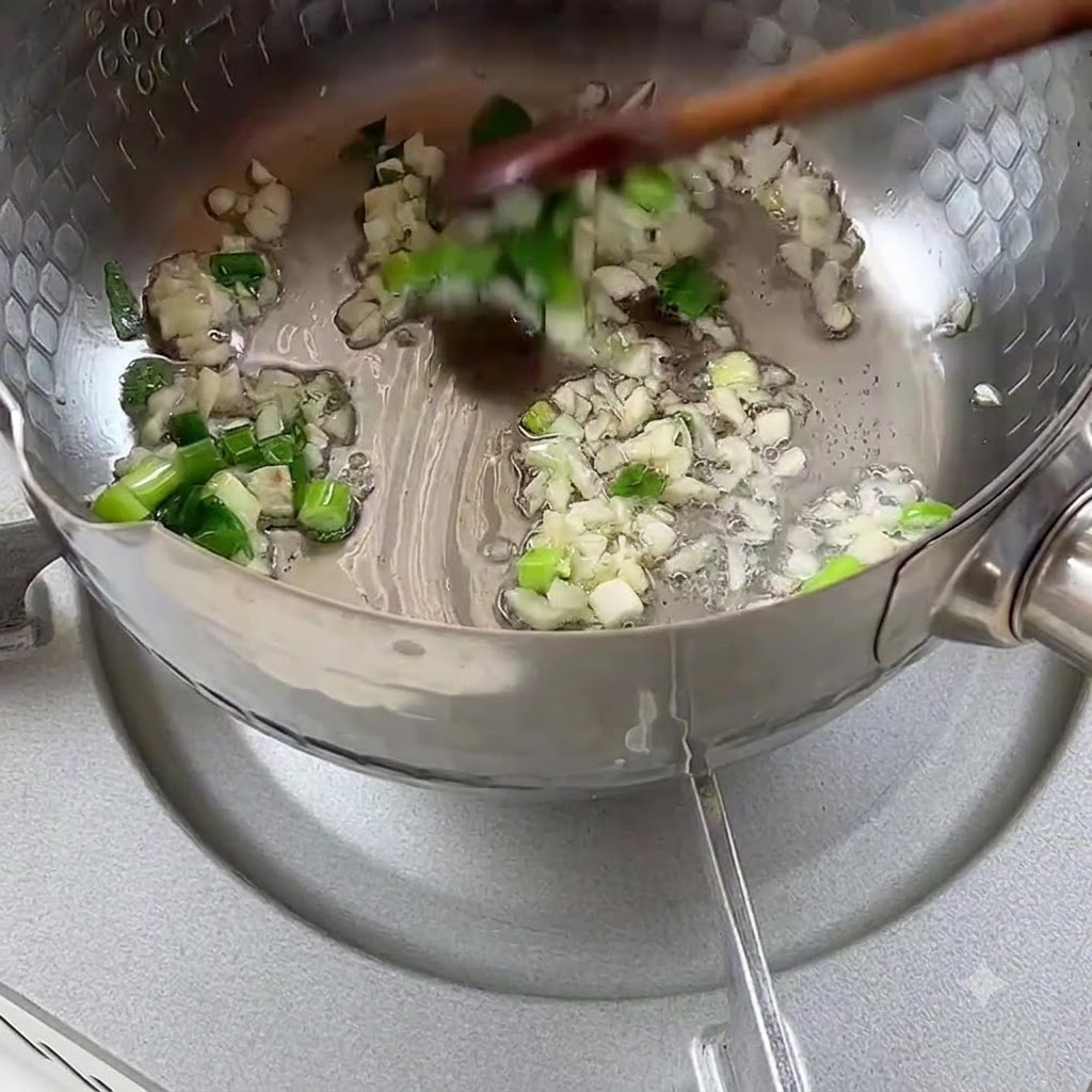 Chopped scallions and garlic sizzling in cooking oil inside a metal pot.
