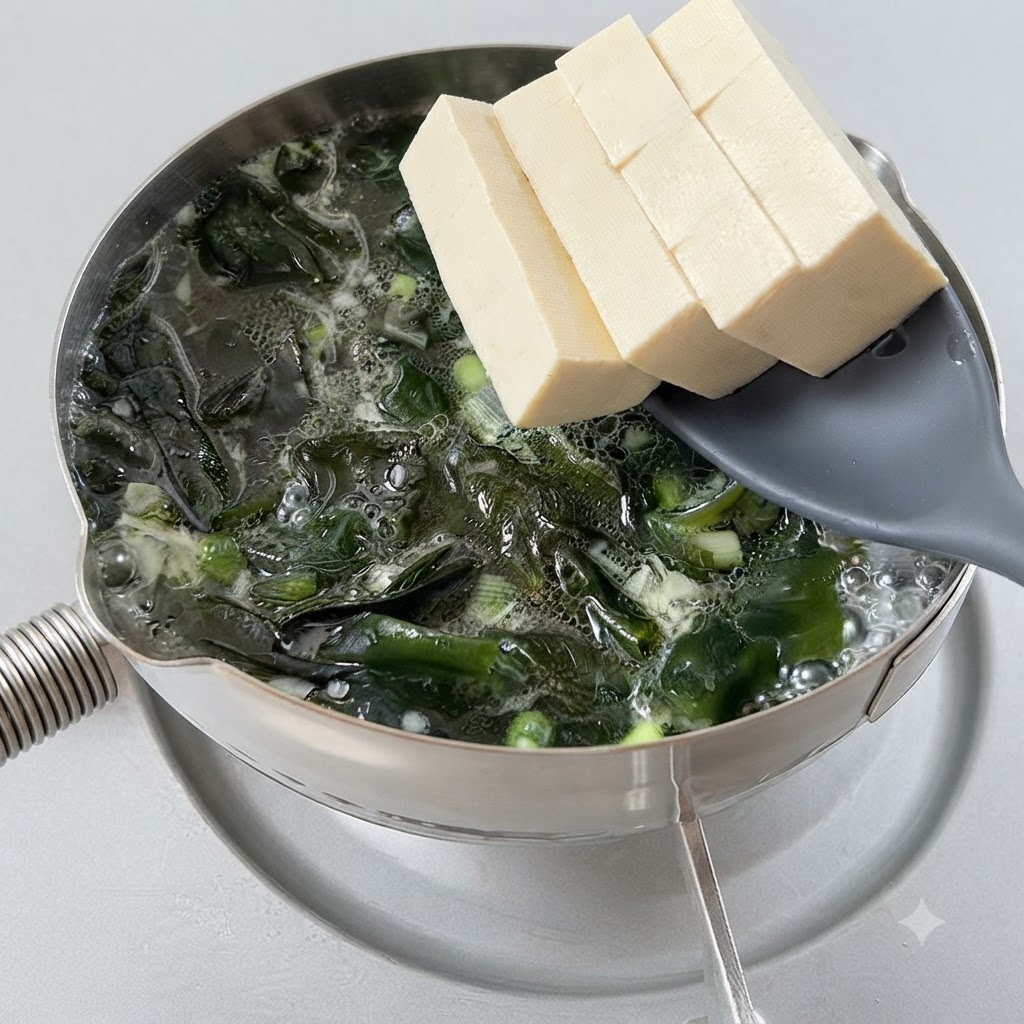 Tofu cubes being transferred into a bubbling pot of seaweed soup using a slotted spoon.